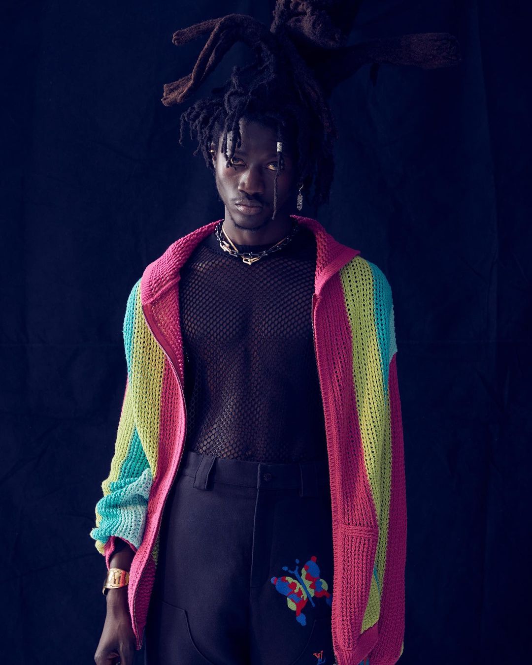 Portrait of an adult male wearing a multicolored sweater, black pants with butterfly designs, and silver jewelry against a dark background.