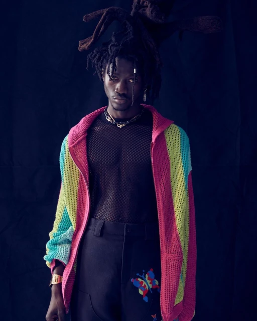 Portrait of an adult male wearing a multicolored sweater, black pants with butterfly designs, and silver jewelry against a dark background.