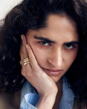 Close-up portrait of a woman resting her face on her hand wearing stacked gold rings and a blue striped shirt