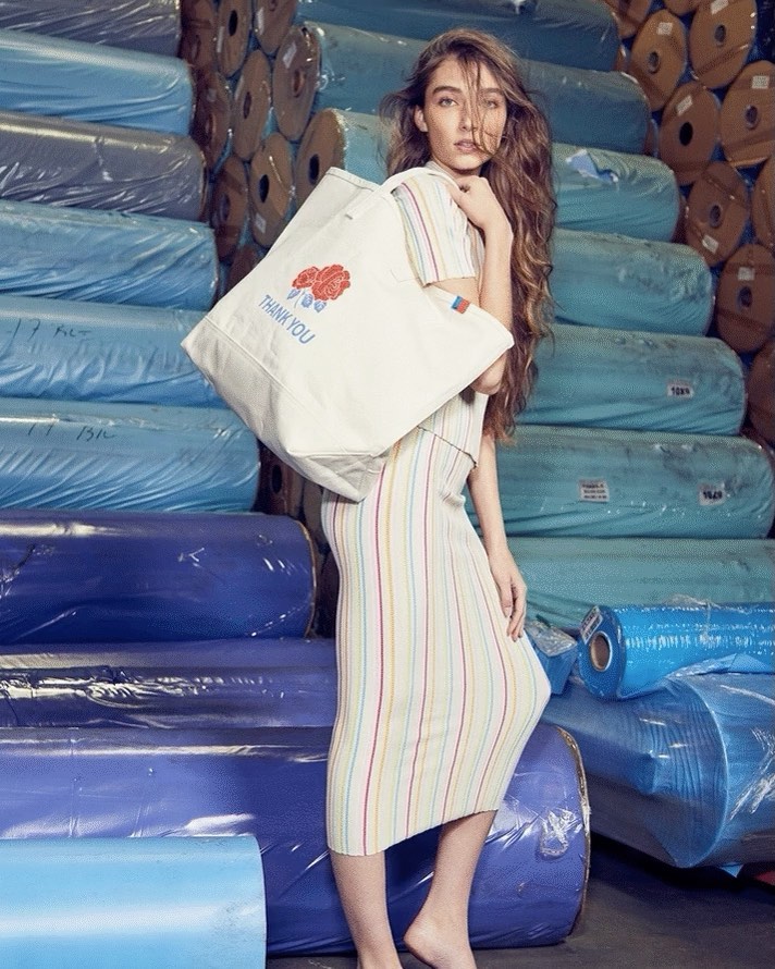 Adult female model poses indoors with a canvas tote bag and striped dress, set against stacked rolls of blue-wrapped textile fabric.
