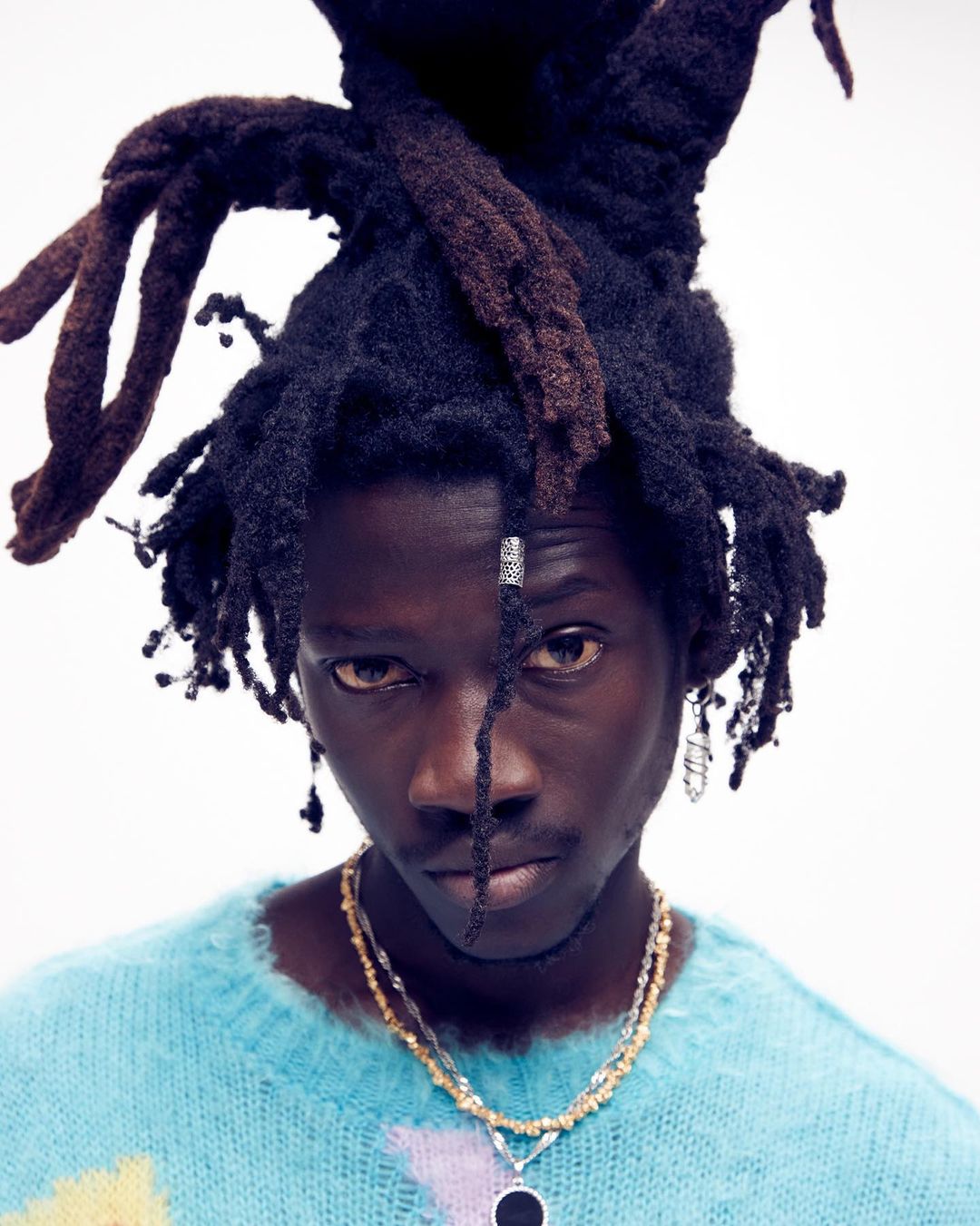 Close-up portrait of a young adult male with dreadlocks wearing a blue and yellow sweater and layered beaded necklaces on a white background.