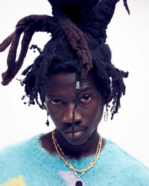 Close-up portrait of a young adult male with dreadlocks wearing a blue and yellow sweater and layered beaded necklaces on a white background.