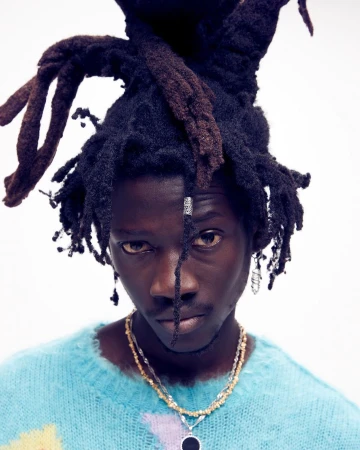 Close-up portrait of a young adult male with dreadlocks wearing a blue and yellow sweater and layered beaded necklaces on a white background.