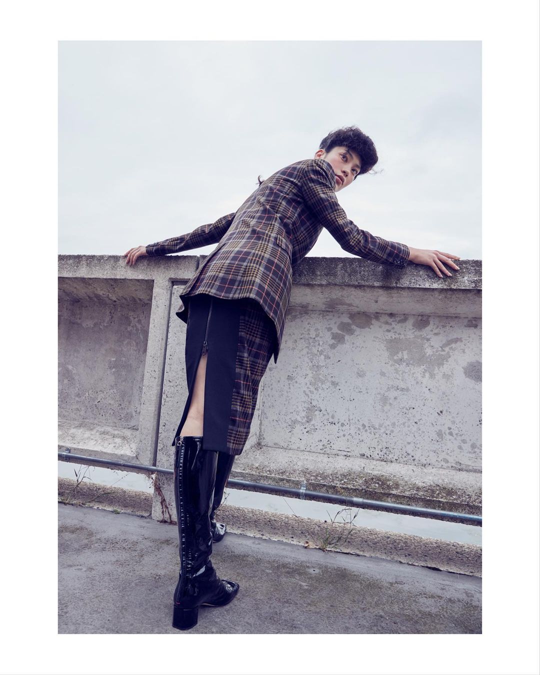 A person leans against a concrete wall wearing a plaid blazer, black skirt, and shiny black boots in an outdoor fashion shot.