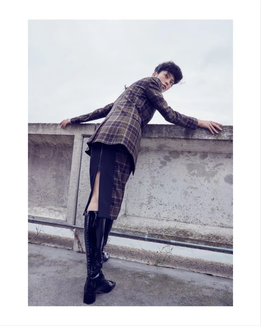 A person leans against a concrete wall wearing a plaid blazer, black skirt, and shiny black boots in an outdoor fashion shot.