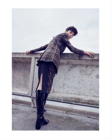 A person leans against a concrete wall wearing a plaid blazer, black skirt, and shiny black boots in an outdoor fashion shot.