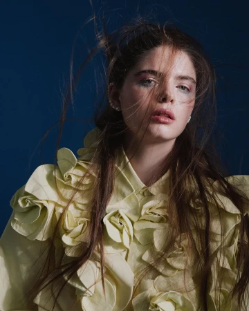 Portrait of a woman with long tousled hair wearing a pale yellow textured floral appliqué shirt against a dark blue background.