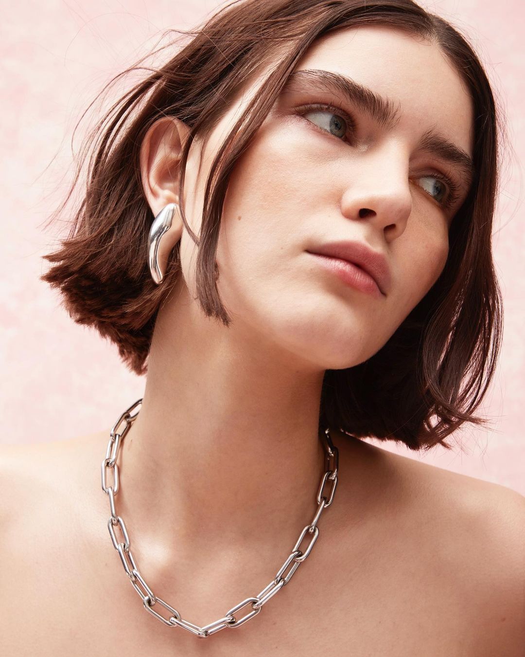 Close-up shot of a woman with short brown hair wearing a silver chain necklace and silver earrings against a pink background.