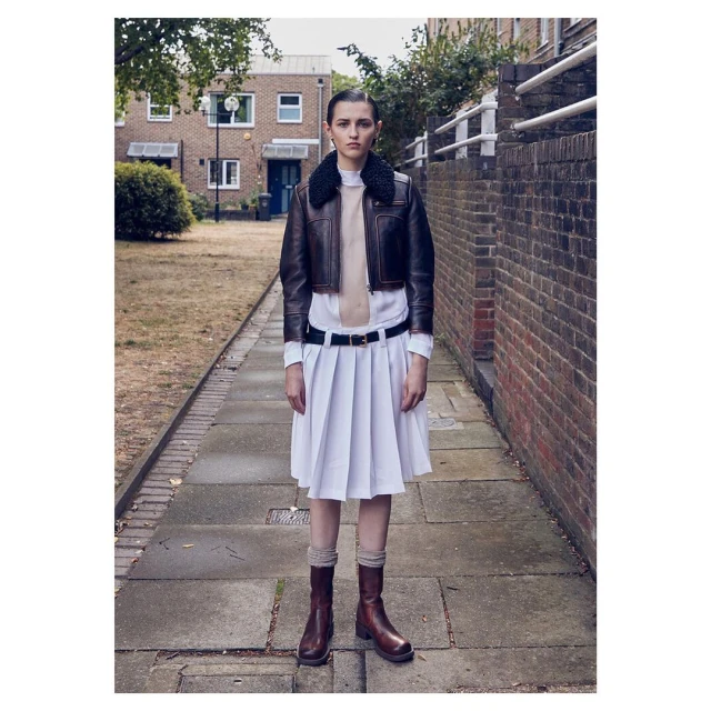 A female adult poses on a brick-paved walkway wearing a brown leather bomber jacket, white dress, brown socks and boots, with brick buildings.