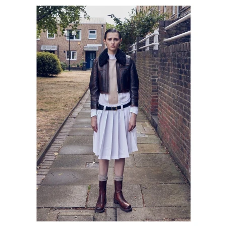 A female adult poses on a brick-paved walkway wearing a brown leather bomber jacket, white dress, brown socks and boots, with brick buildings.