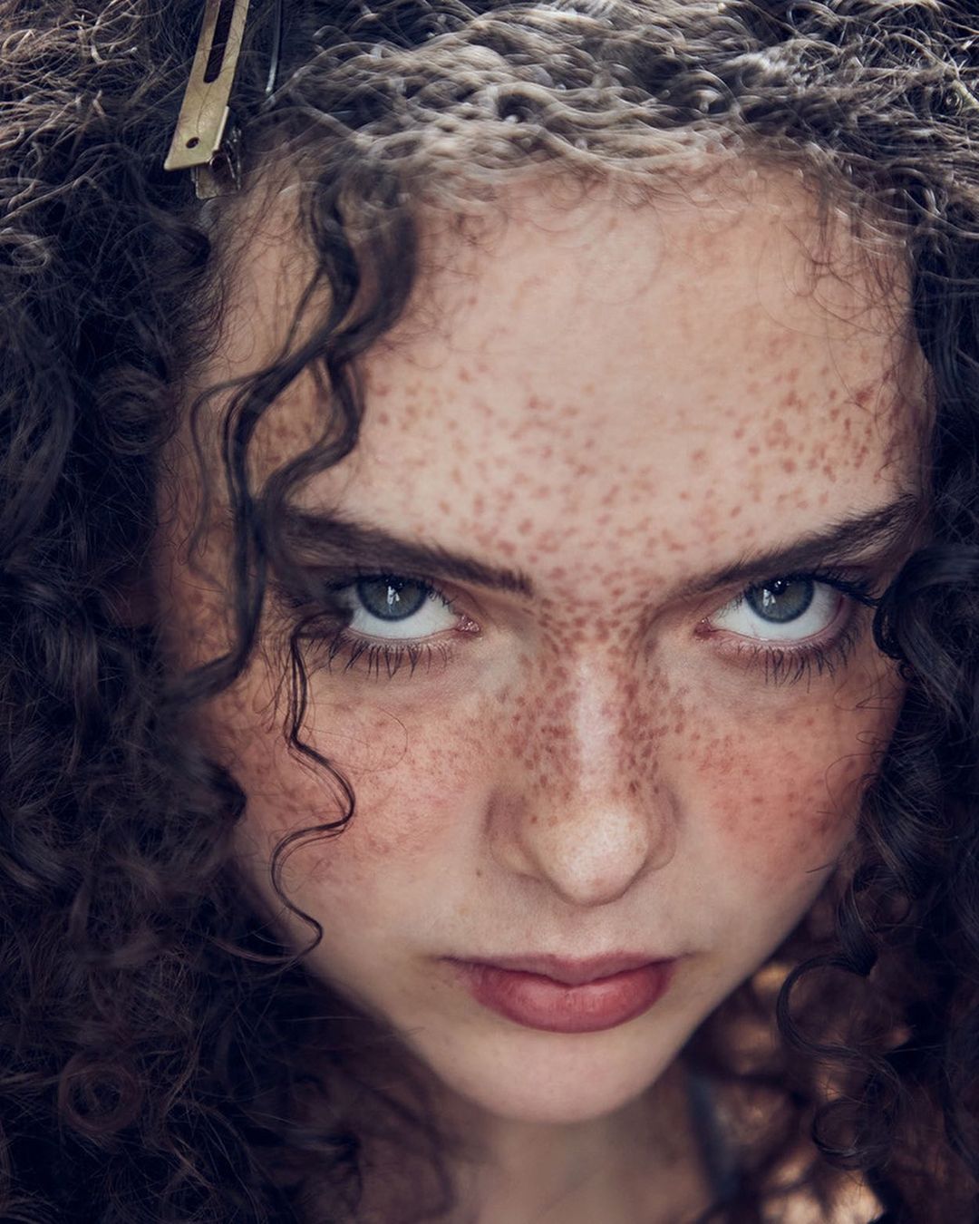 Close-up portrait of a young adult female with freckles, curly hair and a serious expression, wearing a clip in her hair.