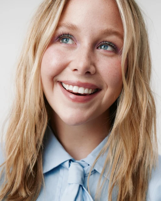 Close-up studio shot of a smiling fair-skinned blonde woman with blue eyes in a light blue collared blouse against a white backdrop.