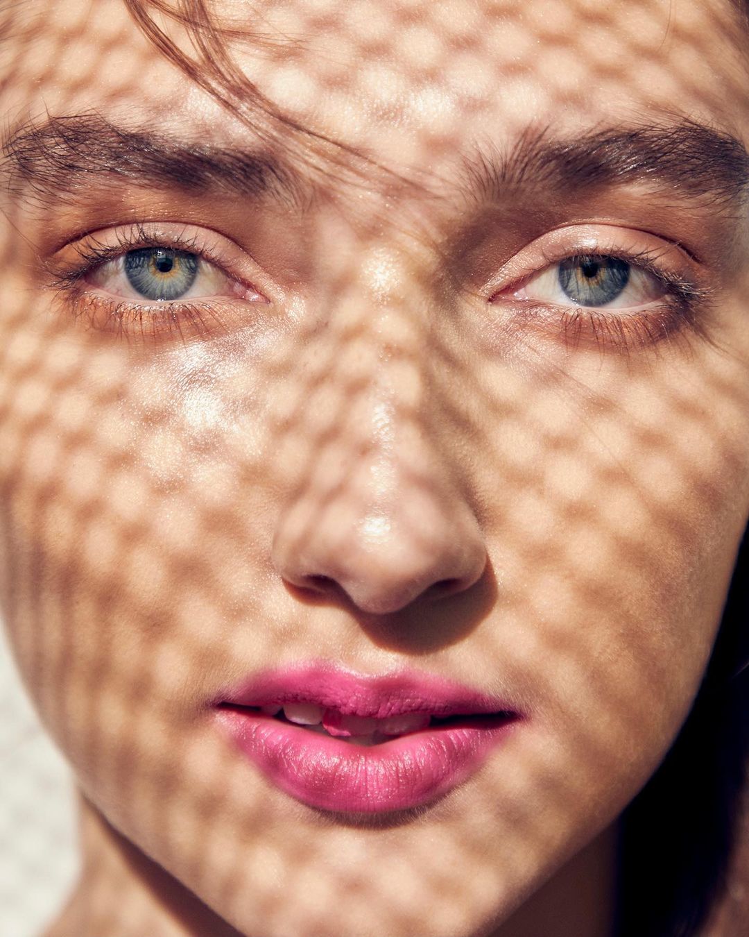 Close-up portrait of a woman with pink lipstick, her face patterned with dappled shadow from an unseen screen or mesh.