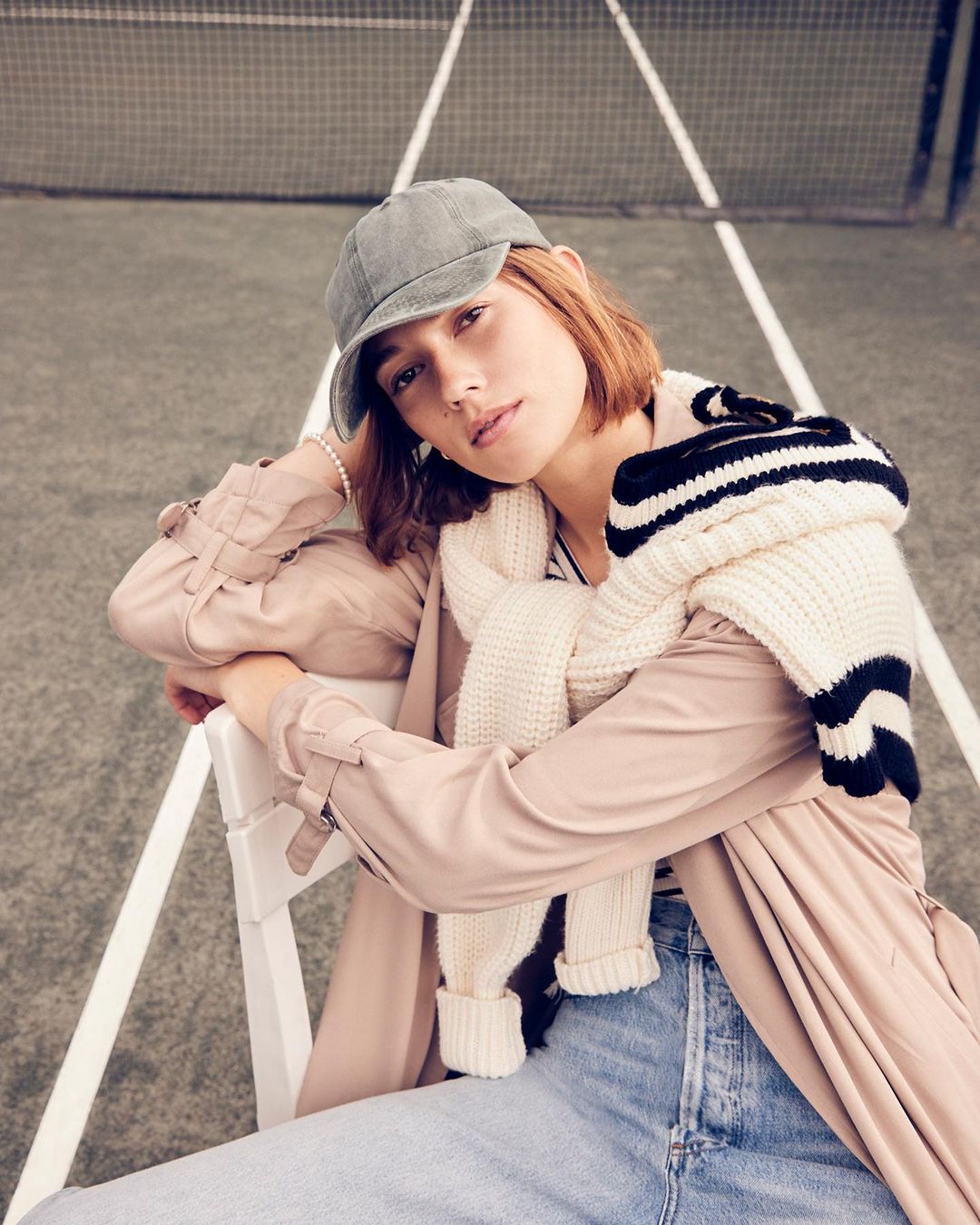A woman in a tan trench coat and baseball cap is seated in front of a grey outdoor court with white lines, fashion portrait.