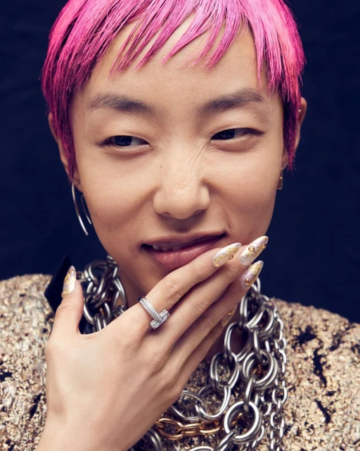 Close-up portrait of an Asian adult female with pink hair, fair skin, wearing a gold top, silver jewelry, and decorative nails on black background.