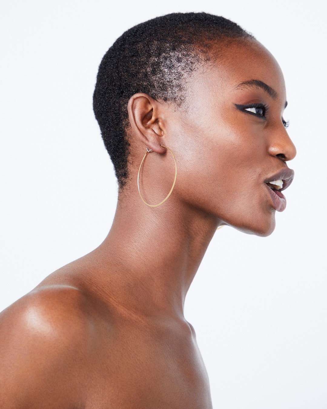 Close-up studio portrait of a woman with an open mouth, showcasing short hair, brown skin, hoop earrings, and eyeliner on a white backdrop.