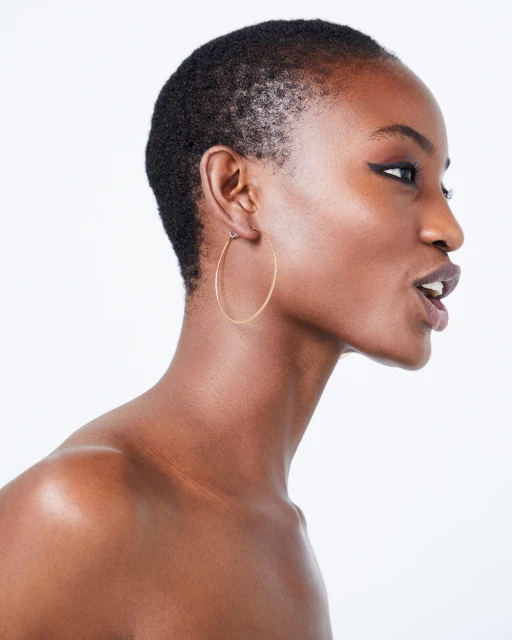 Close-up studio portrait of a woman with an open mouth, showcasing short hair, brown skin, hoop earrings, and eyeliner on a white backdrop.