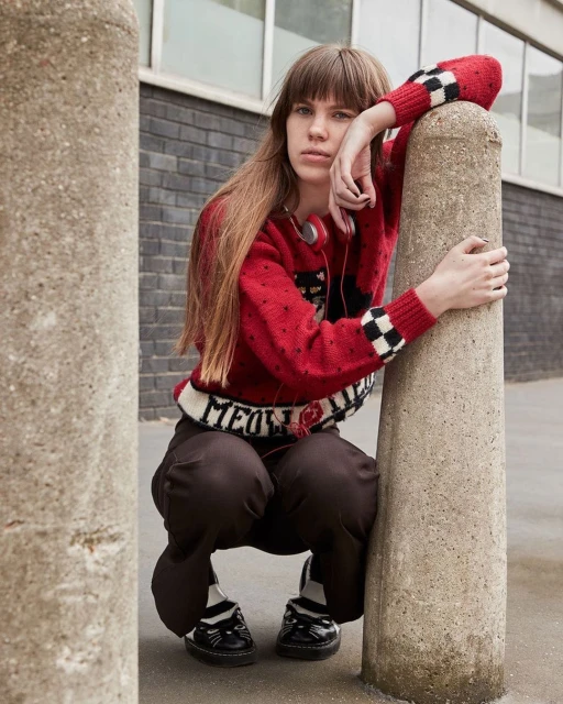 Young woman with long brown hair poses in a red sweater with a ‘Meow’ graphic and dark pants, leaning on a concrete pillar.