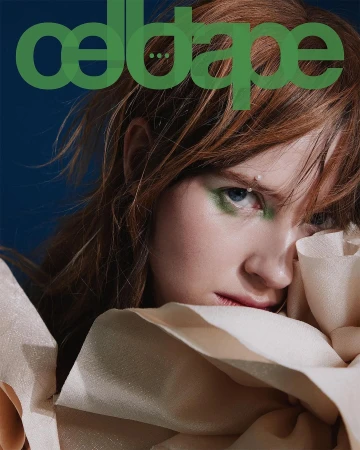 Close-up portrait of a woman with red hair and green eyeshadow, resting her face against textured fabric against a dark blue background.