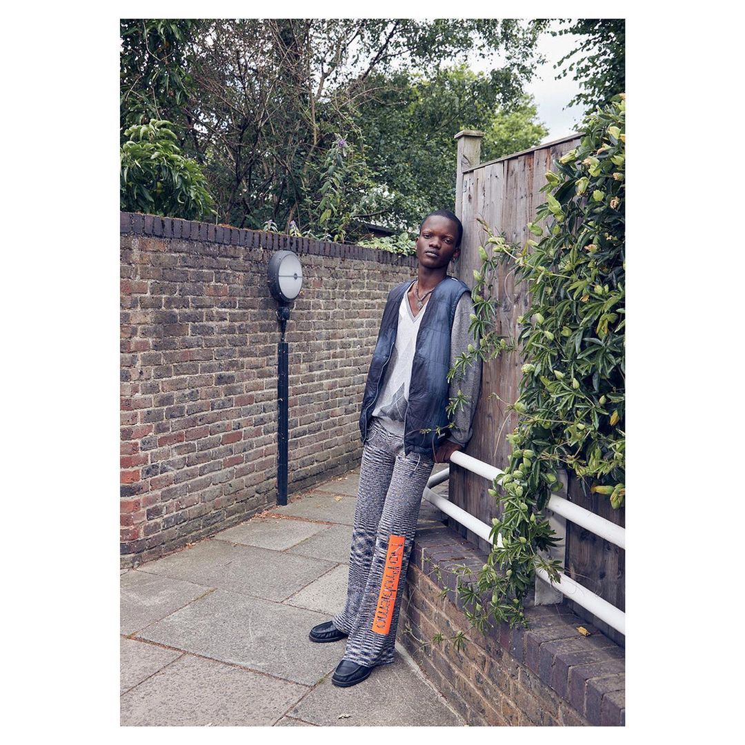Young Black male model wearing gray denim leaning against a fence in an urban outdoor setting with brick walls and greenery.