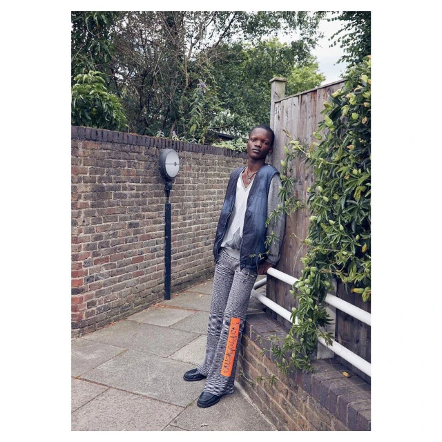 Young Black male model wearing gray denim leaning against a fence in an urban outdoor setting with brick walls and greenery.