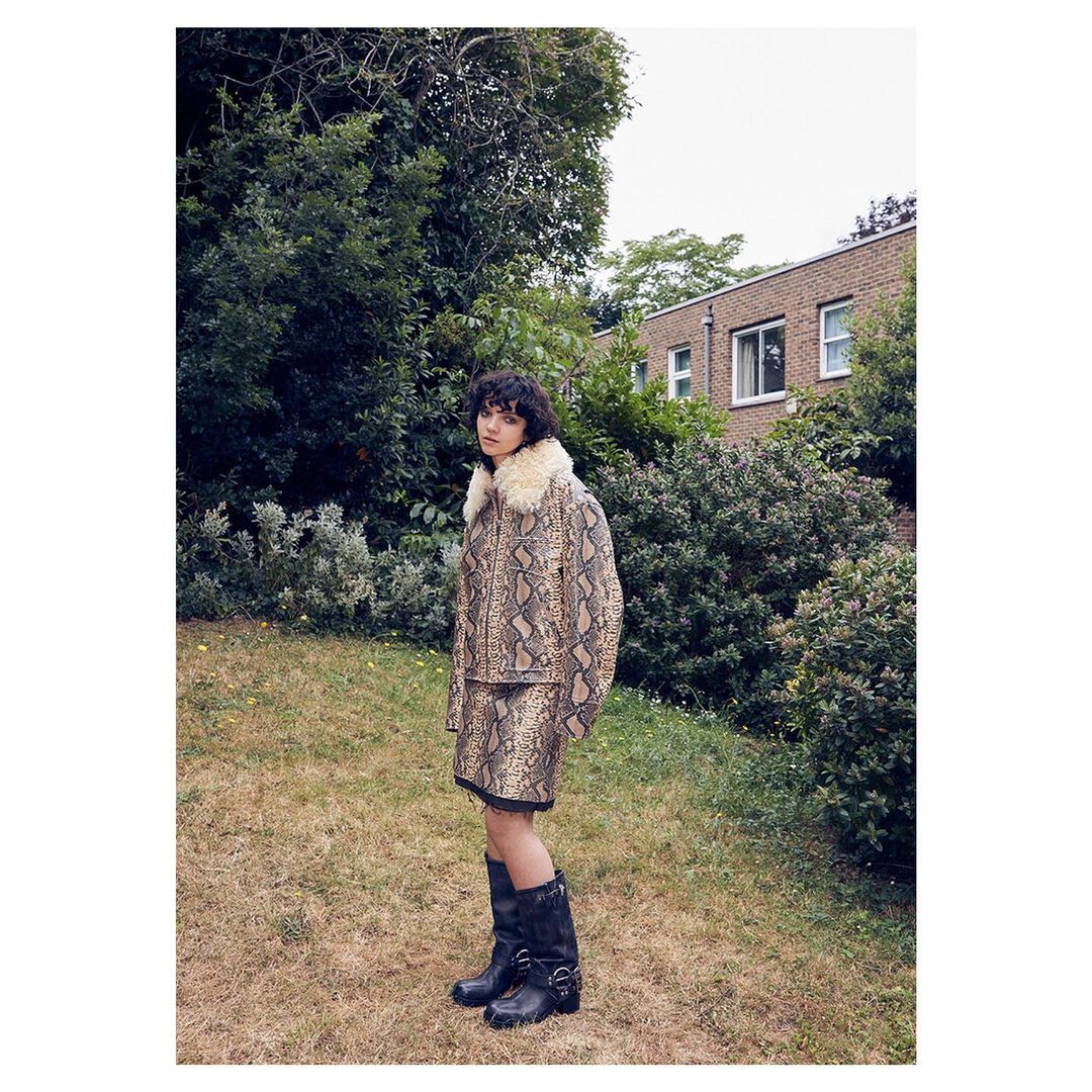 Woman in a snake print jacket and skirt stands outside on a lawn, with trees and a brick building in the background, wearing black boots.