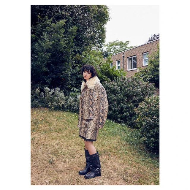 Woman in a snake print jacket and skirt stands outside on a lawn, with trees and a brick building in the background, wearing black boots.
