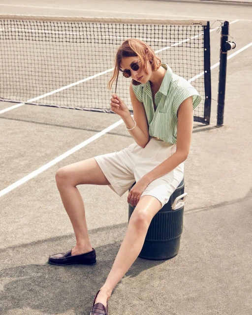 Woman sits on a green cooler on a tennis court, wearing a striped green and white shirt, white shorts, and loafers, with sunglasses.