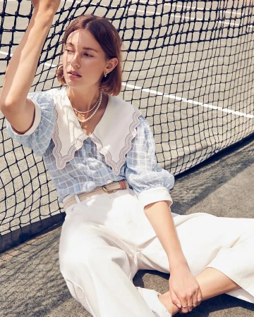 An adult woman with brown hair sitting in a tennis court wearing a blue plaid blouse, white pants, and pearl necklace looking away.