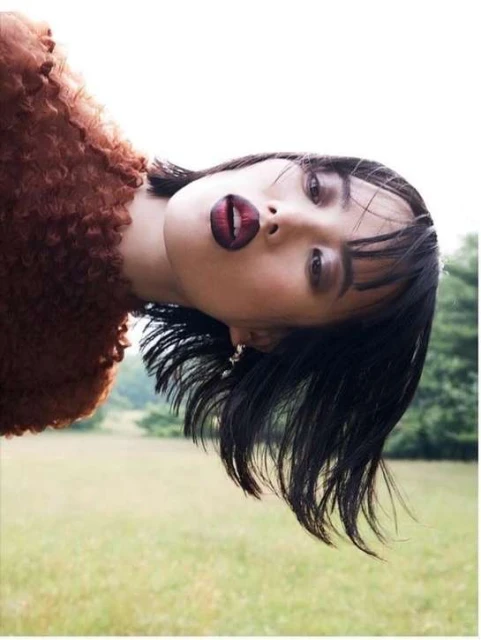 Portrait of a woman with short dark hair wearing a brown textured top, standing in a grassy field with trees in the background.