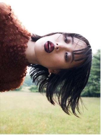 Portrait of a woman with short dark hair wearing a brown textured top, standing in a grassy field with trees in the background.