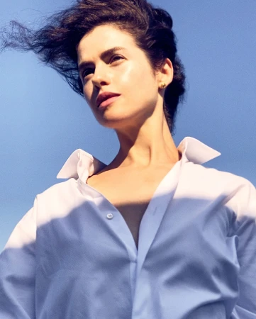 Low angle portrait of a woman with dark hair blowing in the wind wearing a crisp white button-down shirt against a clear blue sky