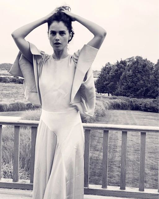 Monochrome portrait of a woman in a gown standing outside, with trees and sky in the background, hands touching her hair.