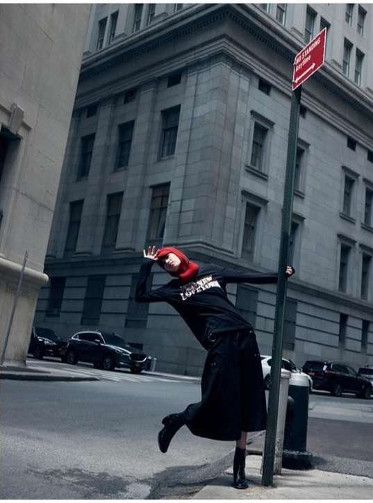 A woman posing on a city street next to a no standing sign, wearing a black top and skirt, with cars and buildings in the background.