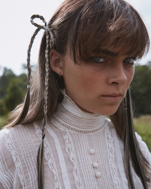 Portrait of a brunette woman with braided pigtails. She wears a white lace blouse against an outdoor backdrop of green grass and trees.