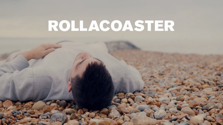 Man in a light shirt lying on a pebble beach with a blurred ocean background and text overlay reading 'ROLLACOASTER'.
