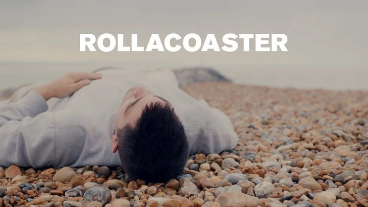 Man in a light shirt lying on a pebble beach with a blurred ocean background and text overlay reading 'ROLLACOASTER'.