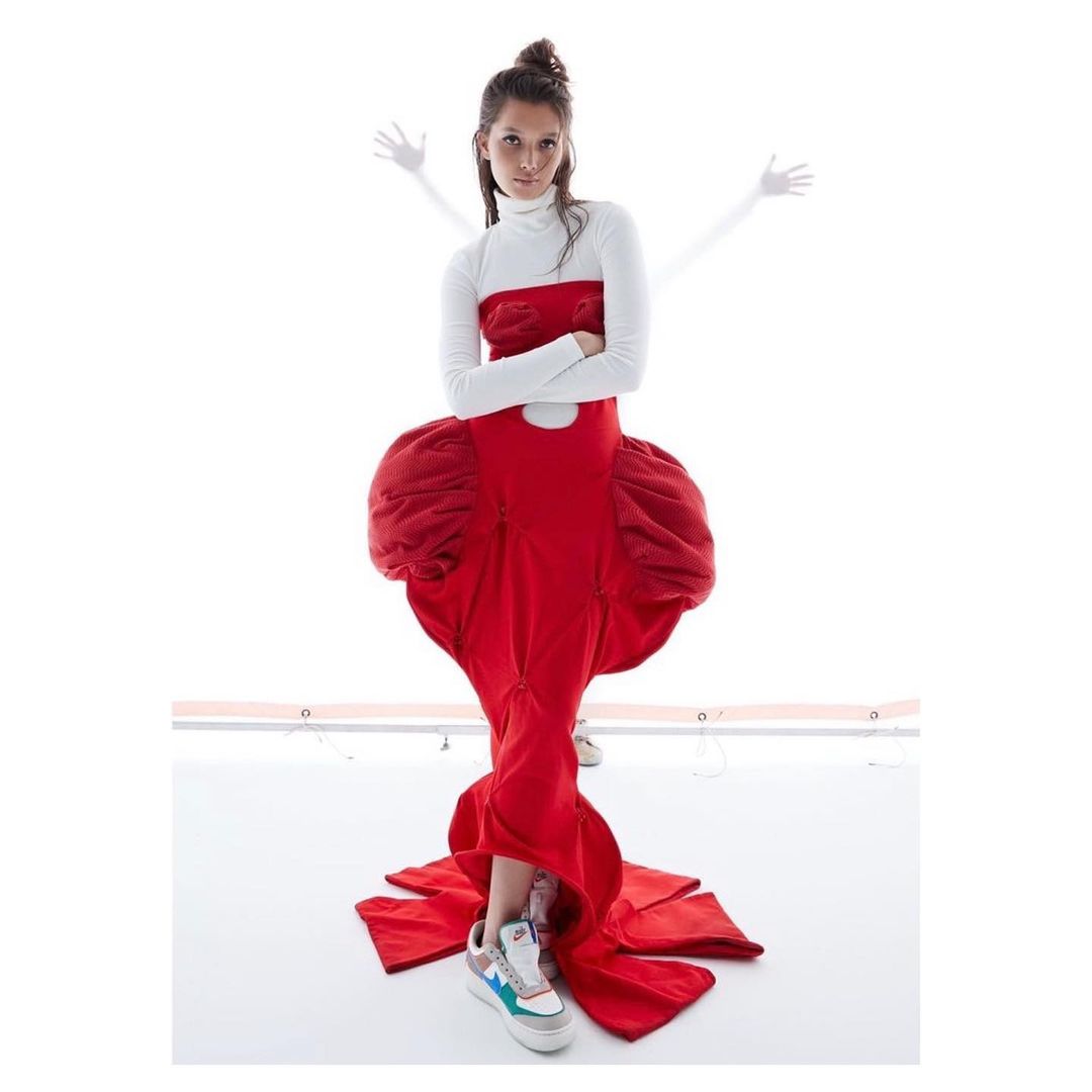 A model poses in a red gown with exaggerated hips against a stark white background with diffused lighting and ghost-like arms in the background.