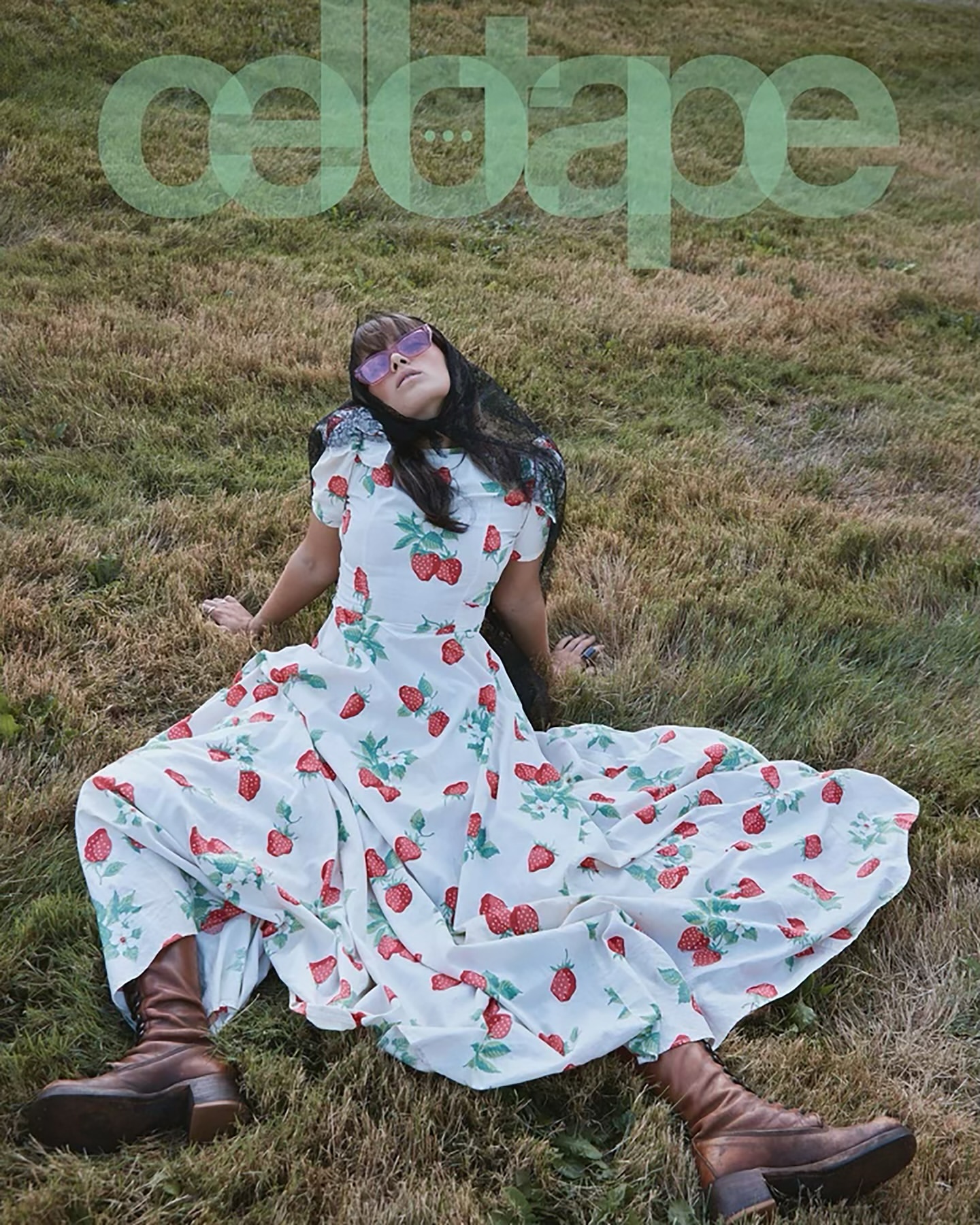 Woman in strawberry-print dress lounges in grassy field wearing sunglasses and brown boots under transparent overlay of ODDA magazine title.