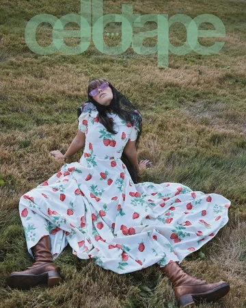 Woman in strawberry-print dress lounges in grassy field wearing sunglasses and brown boots under transparent overlay of ODDA magazine title.