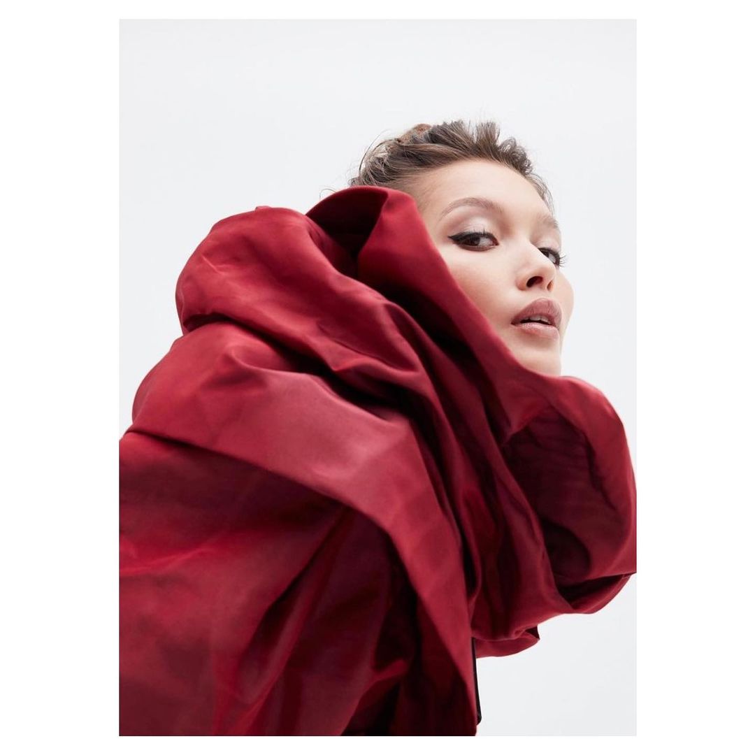 Model wearing a voluminous crimson garment wrapped around the upper body against a plain white background