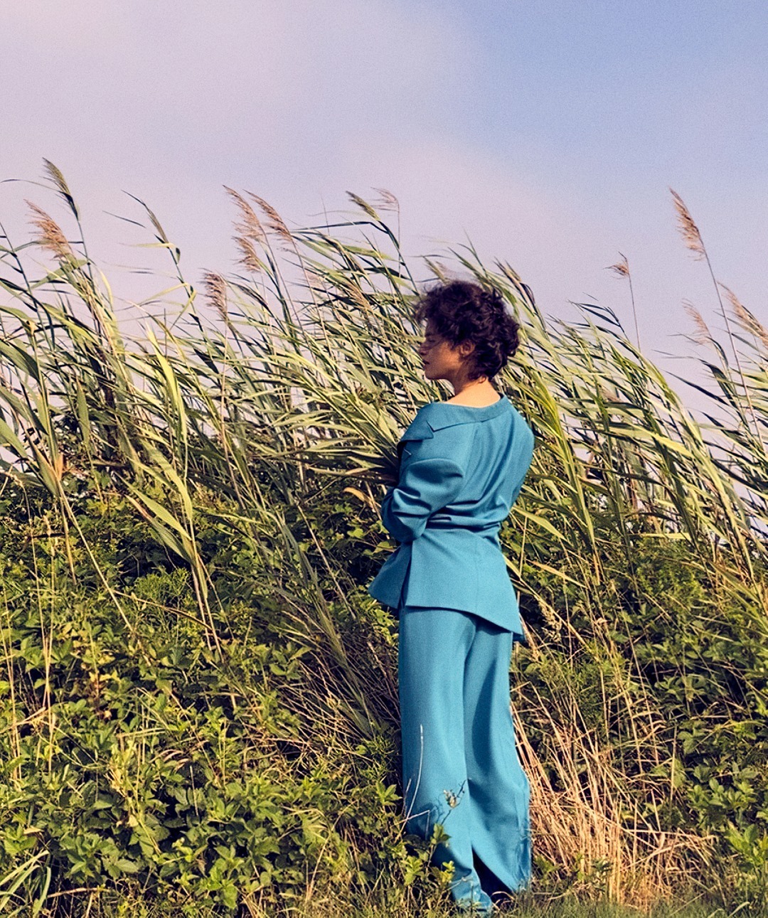 A woman in a blue suit standing in a field of tall grass and reeds against a pastel sky, showcasing fashion in a natural setting.