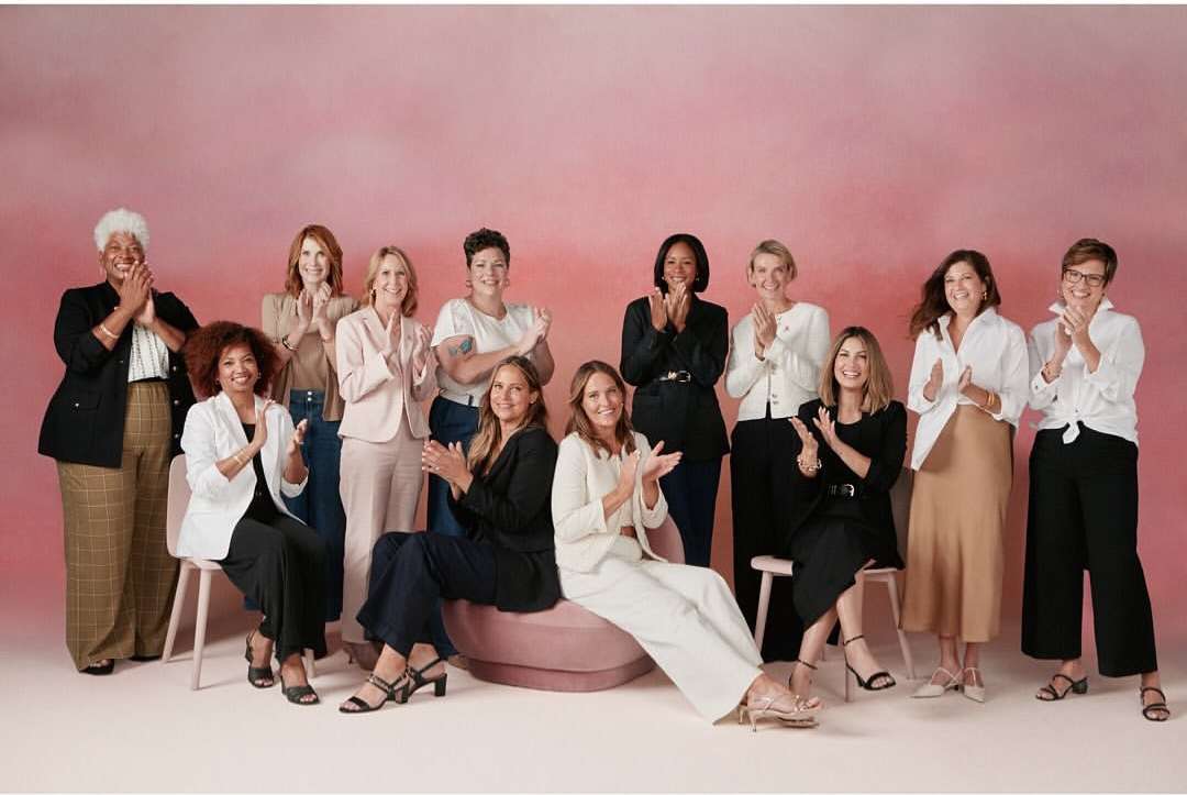 Group of women in business casual attire stand and sit in a studio, applauding against a pink backdrop, capturing a collaborative and celebratory moment.