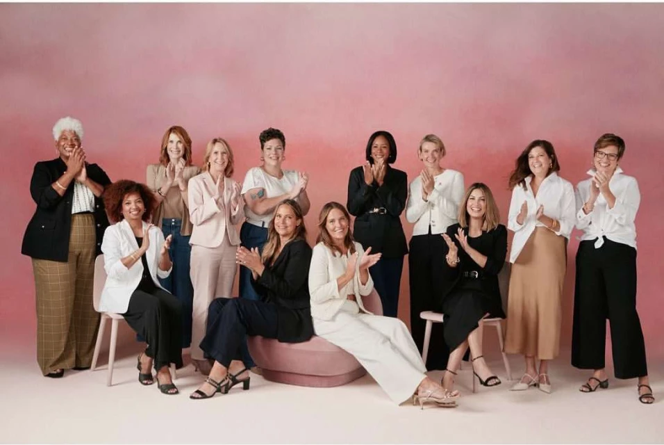Group of women in business casual attire stand and sit in a studio, applauding against a pink backdrop, capturing a collaborative and celebratory moment.