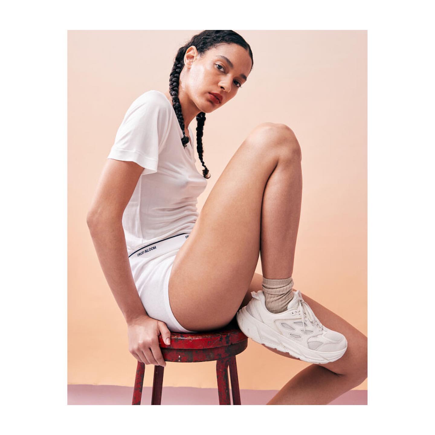 An adult woman with dark hair in braids, wearing white athletic clothing, poses indoors while sitting on a red stool in a studio.