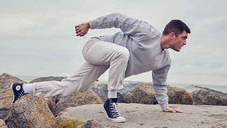 Adult male in athletic pose on rocky beach wearing grey sweatshirt, white pants and black high-top sneakers against cloudy sky.