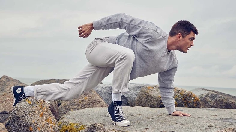 Adult male in athletic pose on rocky beach wearing grey sweatshirt, white pants and black high-top sneakers against cloudy sky.