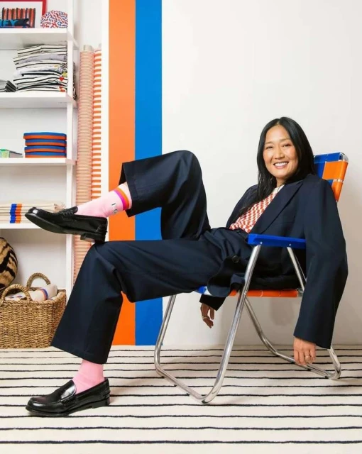 Stylish Asian woman in oversized suit smiles while seated in a chair, wearing pink socks and loafers, with a striped carpet.