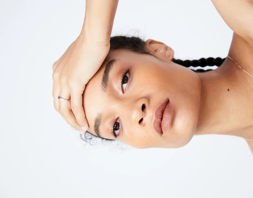 Close-up portrait of a young woman with brown skin and braided hair against a white background, looking directly at the camera.