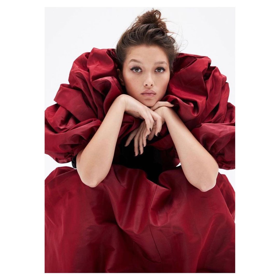 Portrait of an Asian woman wearing a red ruffled dress, leaning on her forearms with a neutral facial expression, against a white backdrop.
